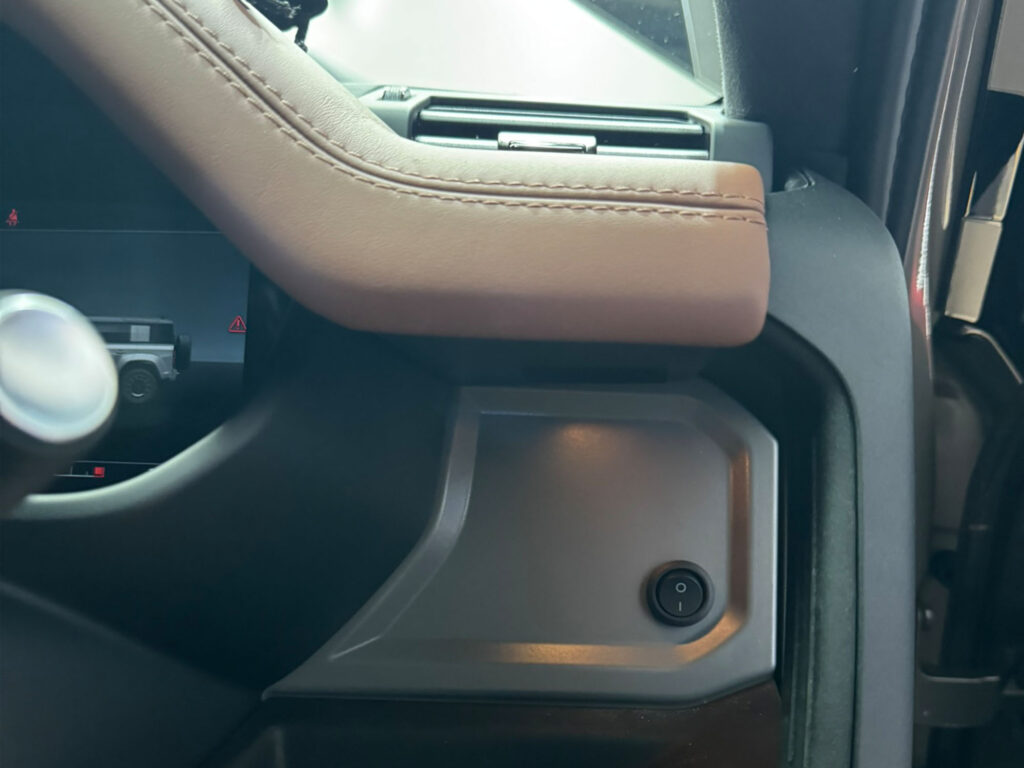 Close-up detail of the custom-installed rocker switch for the front grille lightbar, mounted flush within the Land Rover Defender's factory dashboard trim for an original equipment manufacturer style finish.